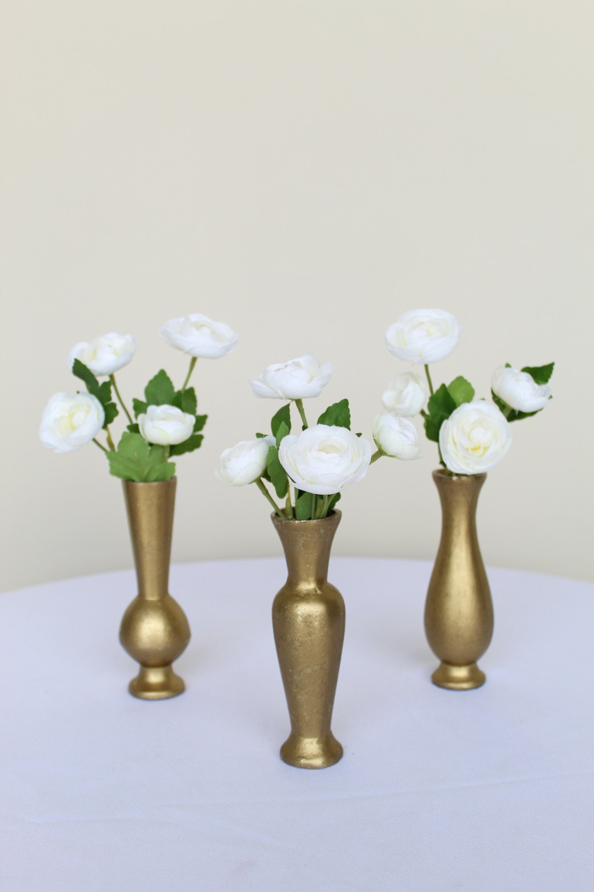 White ranunculus bud vase flowers inside three gold bud vases on a white table in front of a white backdrop