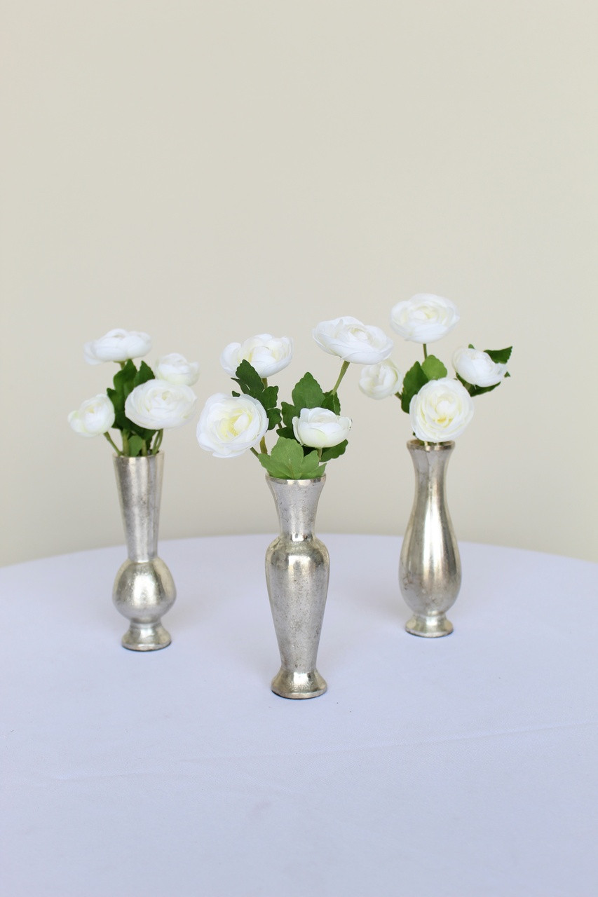 White ranunculus bud vase flowers inside three silver bud vases on a white table in front of a white backdrop