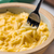 A close up image of a skillet of It's Skinny mac and cheese, with a silver forked dipped into the cheese low-carb pasta.