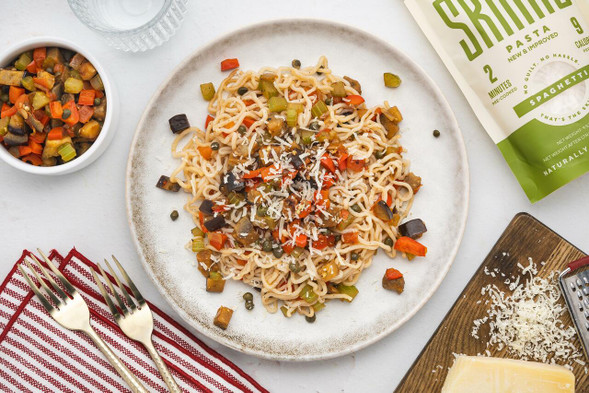 An overhead image of eggplant caponata with It's Skinny spaghetti served on a white plate. A small bowl of sautéed vegetables, grated Parmesan cheese, and a package of It's Skinny spaghetti are arranged around the dish.
