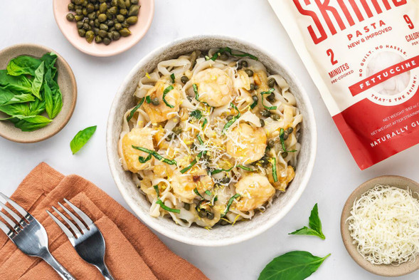 An overhead image of a bowl of It's Skinny Shrimp Piccata. Around the bowl are smaller bowls of ingredients -- Parmesan cheese, fresh basil, capers, and a bag of It's Skinny Fettuccine pasta.