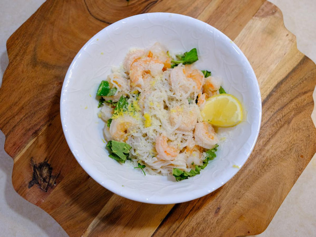 An overhead image of a while bowl filled with pasta and shrimp in a creamy coconut sauce. The dish is garnished with fresh parsley and lemon wedges.