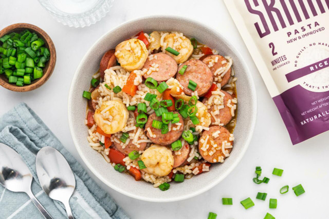 An overhead image of a Cajun rice bowl made with seasoned shrimp, andouille sausage, veggies, and It's Skinny rice. Around the bowl is a set of silverware, a bag of rice, and a small bowl of sliced green onions.