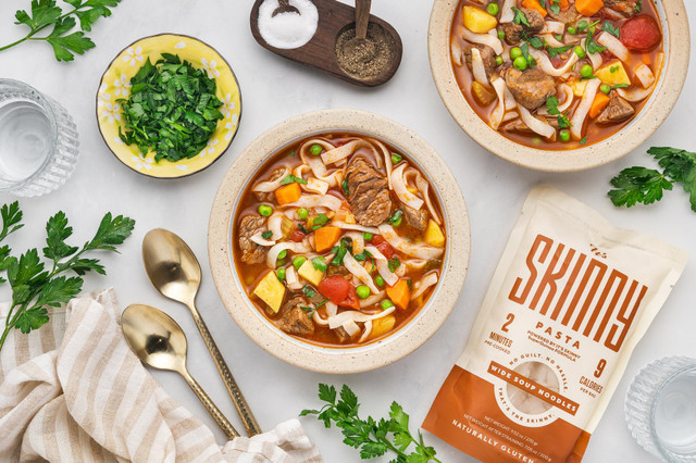 Two bowls of heart beef vegetable soup served at a table filled with flatware, napkins, and small dishes of herbs and spices. A bag of It's Skinny soup noodles rest next to the full bowls.