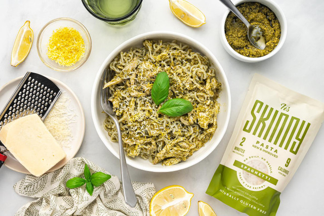 An overhead image of a bowl of chicken pesto spaghetti. Around the bowl are recipe ingredients -- Parmesan cheese, lemon wedges, pesto sauce, and a bag of It's Skinny spaghetti.
