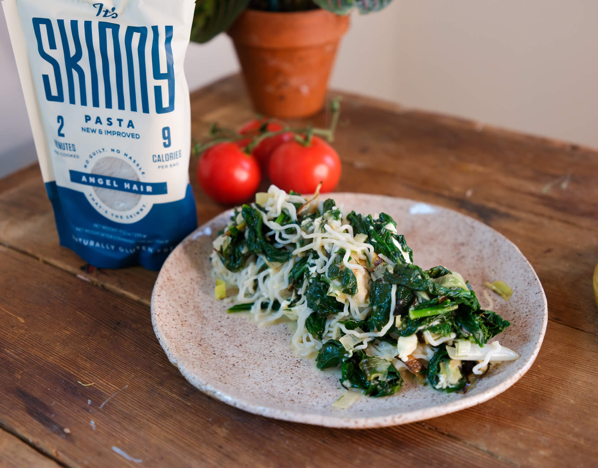 A white plate full of spinach and feta pasta sits on a wooden tabletop, next to a bag of It's Skinny angel hair pasta.