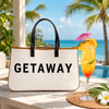White canvas tote bag printed with "GETAWAY" in bold black text, featuring black leather handles and brown trim.