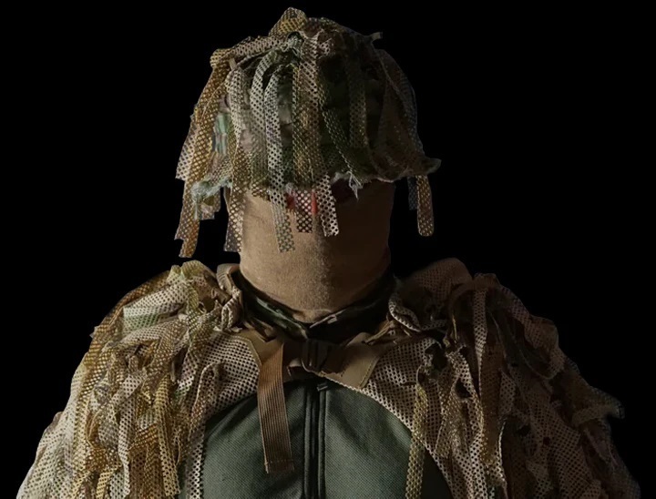 An operator wearing Carcajou sniper boonie hat attach with extra boonie garnish, also wearing a coyote brown balaclava and badger, behind the operator is a black background.