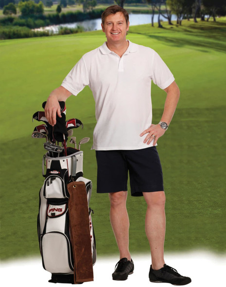 A man stands by a golf bag wearing navy chino shorts and a white polo shirt, with a lush green landscape in the background.