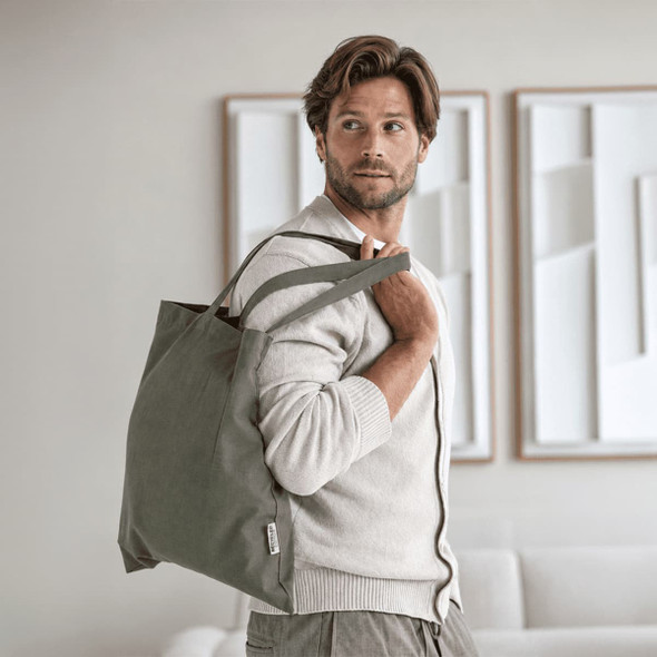 A man carries a large grey tote bag, wearing a light sweater, in a modern interior setting. The bag has a logo.