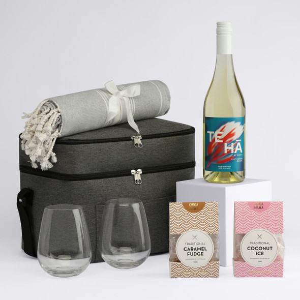 A drink bottle and two stemless glasses sit beside a picnic basket, blanket, and two gourmet treats in pink packaging.