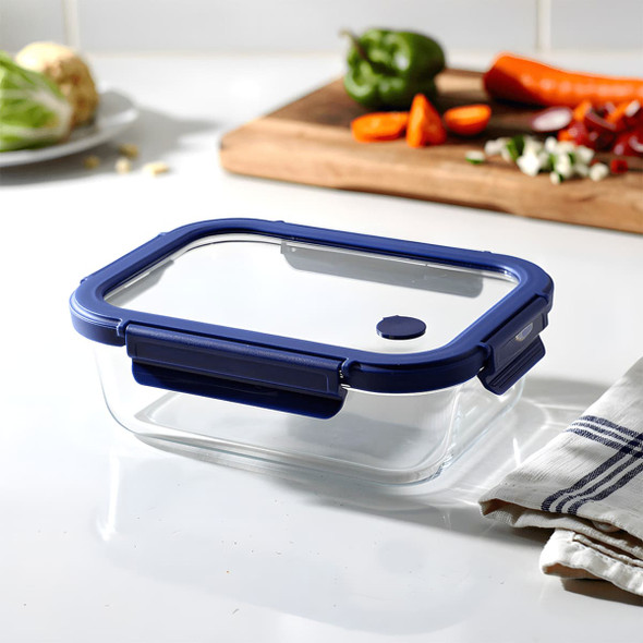 A glass lunch box with a navy blue plastic lid, placed on a kitchen counter next to fresh vegetables and a towel.