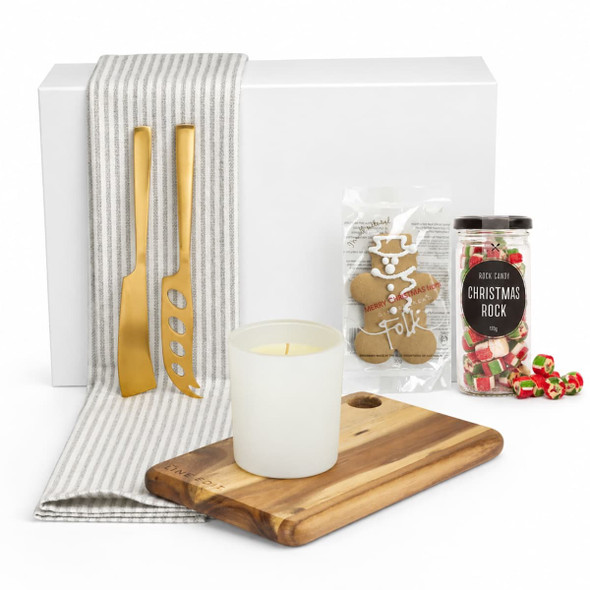A white box contains a wooden serving board, gold utensils, a candle, gingerbread, and Christmas sweets, all on a striped cloth.