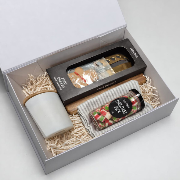 A festive hamper featuring a candle, a cheese knife set, and a jar of Christmas treats, all in a white gift box.