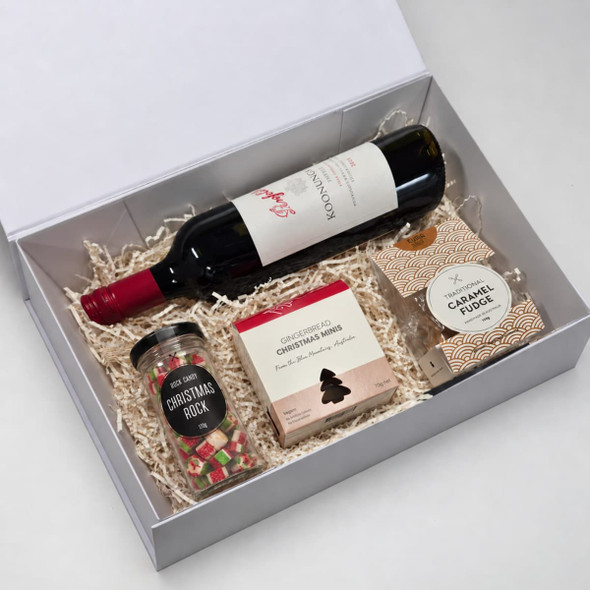 A gift box containing a red wine bottle, Christmas rock sweets, Christmas mix chocolate, and caramel fudge on shredded paper.