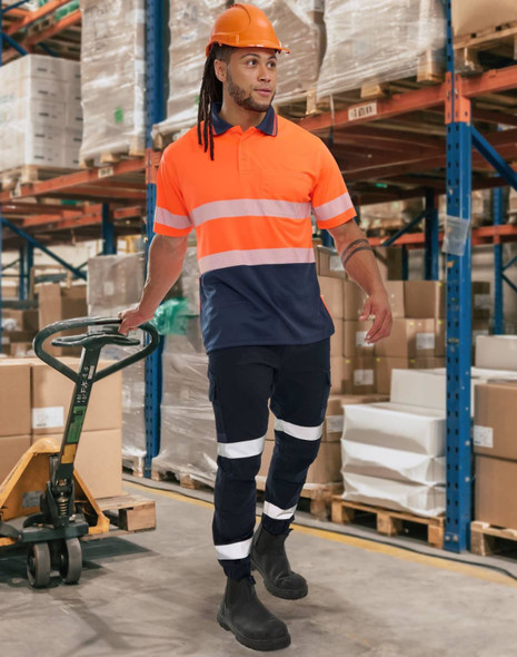 A man wearing orange and navy work pants with reflective tapes, accompanied by a hard hat and boots, stands in a warehouse.