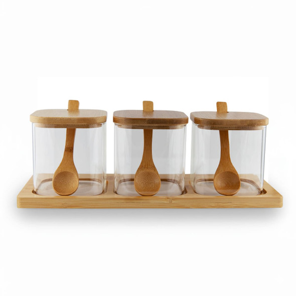 Three glass condiment jars with bamboo lids and wooden spoons, placed on a bamboo tray.