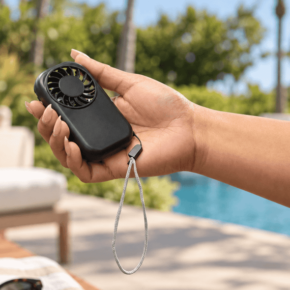 A handheld black mini pocket fan with a wrist strap, being held outdoors near a pool.