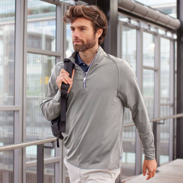 A man wearing a light grey half-zip sweatshirt, carrying a shoulder bag, walking in a modern building.