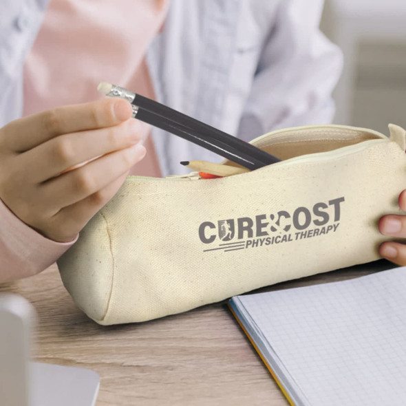 A cream pencil case with a logo, containing pencils and a pen, placed on a wooden surface next to a notebook.