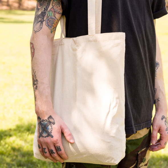 A person holds a natural calico long handle tote bag, featuring straps and a simple design, set against a grassy background.