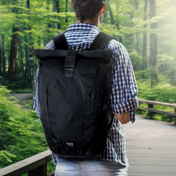 A black roll-up computer backpack with a logo, worn by a person in a checked shirt, against a green forest backdrop.