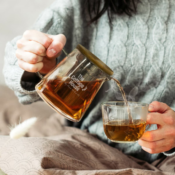 A glass teapot with a wooden lid pours hot tea into a clear cup, set on a soft blanket.