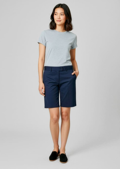 A woman stands wearing navy dress shorts and a light grey T-shirt, with her hands in the pockets. The image features a logo.