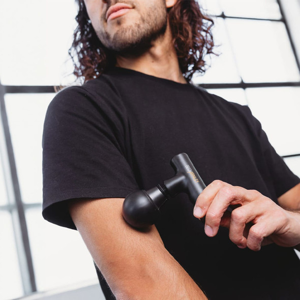 A black mini massage gun is being used on a person's arm. The setting features large windows with natural light.