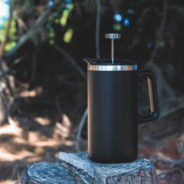 A black coffee press with a stainless steel lid and handle, resting on a wooden stump in a forested setting.