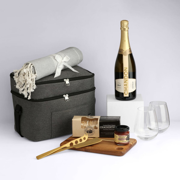 A grey hamper contains a bottle of sparkling wine, two glasses, a cheese board, a blanket, and gourmet snacks.