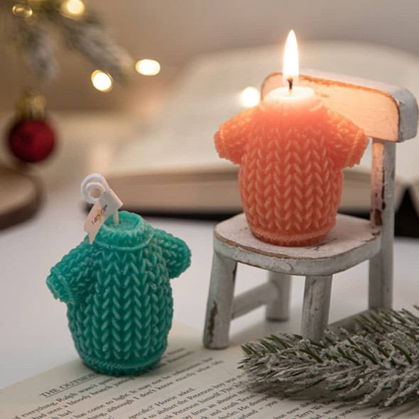 Two sweater-shaped candles in teal and orange, sitting on a small chair with soft lighting and a blurred background.