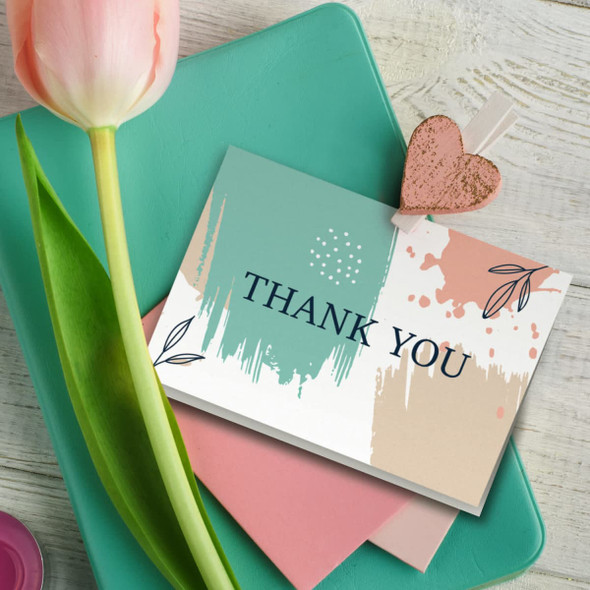 A thank you card with a teal and peach abstract design, resting on a green surface with a pink envelope and a tulip.