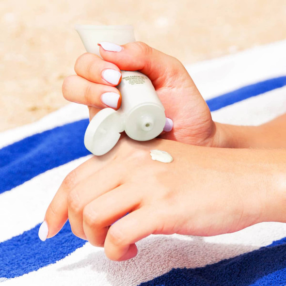 A hand applying white sunscreen from a 50ml tube onto a wrist, set against a beach towel with blue and white stripes. The tube has a logo.