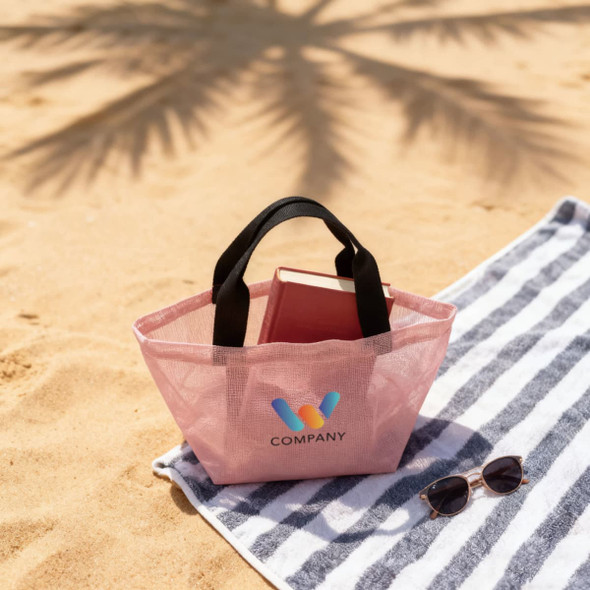 A pink mesh tote bag with black handles rests on a striped beach towel, accompanied by sunglasses and a book.