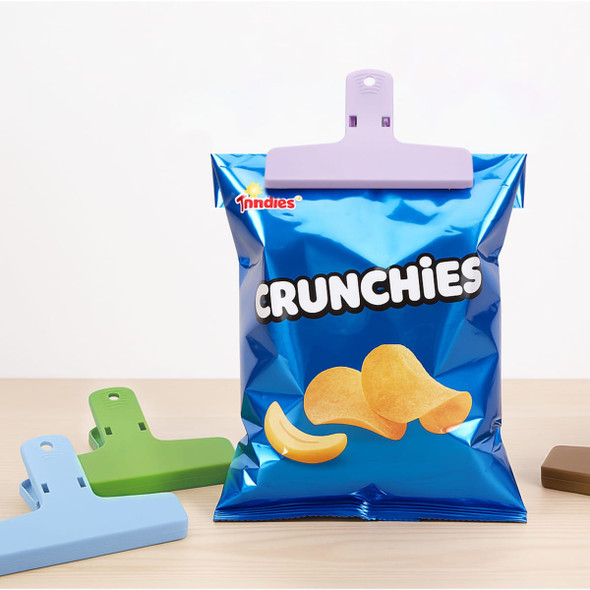 A bag of blue snack chips with a purple plastic clip, accompanied by green and brown plastic clips on a wooden surface.