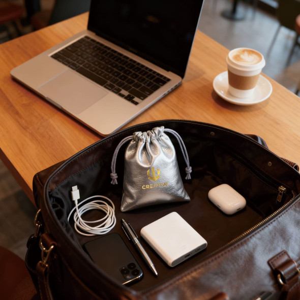 A silver PU leather drawstring gift bag inside a brown bag, with a laptop, phone, charger, and coffee on a table.