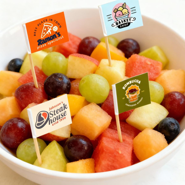 A bowl of mixed fruit featuring watermelon, grapes, and melon, decorated with branding flag sticks.