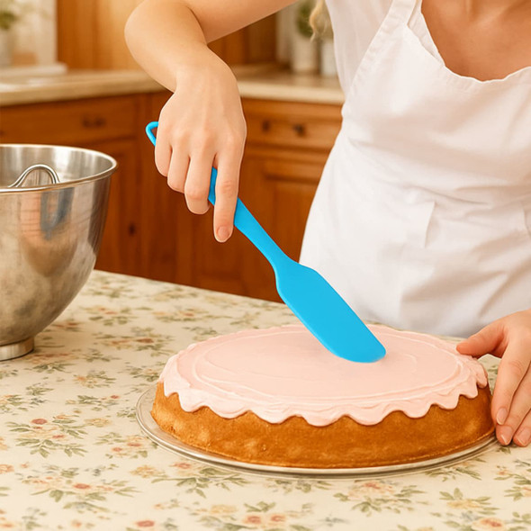 A blue spatula is being used to spread icing on a cake, set on a floral tablecloth with a mixing bowl nearby.