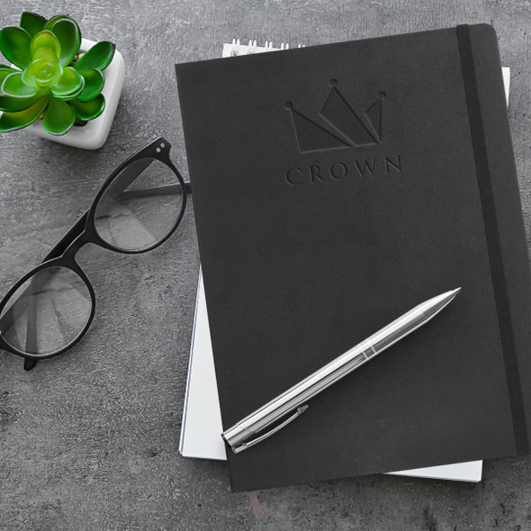 A black Moleskine Classic Hard Cover Notebook with a pen and glasses, placed on a textured surface with a plant.