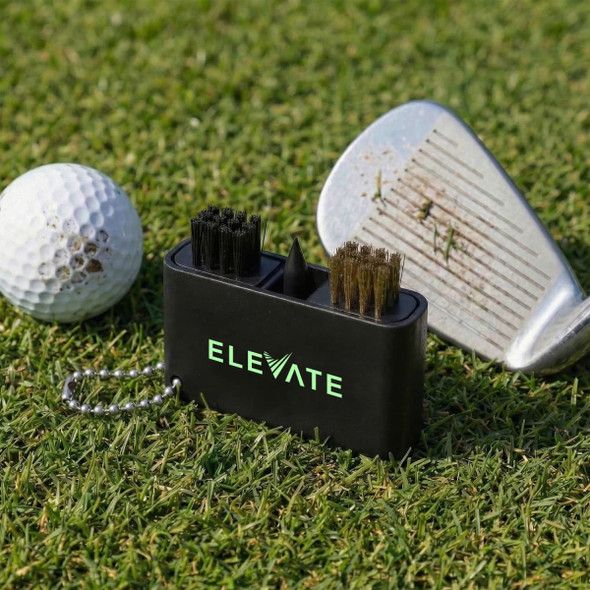 A black golf club cleaning tool with brushes sits on grass next to a golf ball and a club with dirt on it.