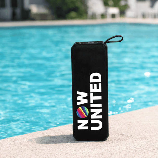 A black portable speaker with a round logo, sitting by a blue pool.