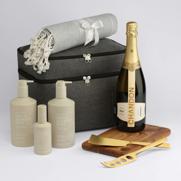 A grey storage bag contains a throw blanket, champagne bottle, and skincare products in elegant packaging on a wooden tray with serving tools.