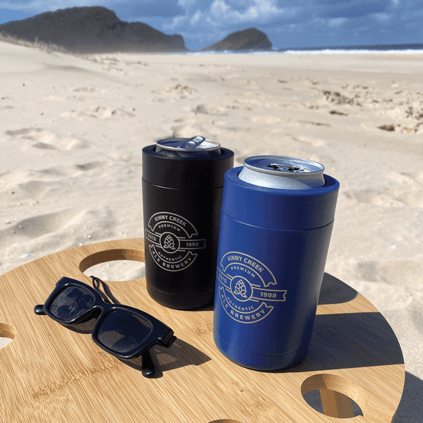 Two stubby holders in black and blue, holding cans, set on a wooden table on a beach. Appropriate for outdoor use.