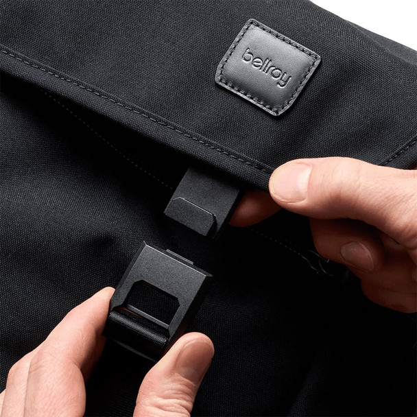 A black messenger bag with a flap and fastening mechanism, featuring a logo. A hand is holding the clasp.