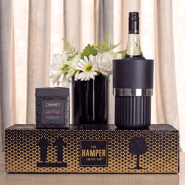 A drink bottle in a black cooler, alongside a box of brittle and a black vase with white flowers.