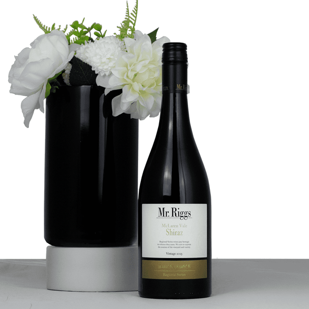 A black wine bottle of Mr. Riggs McLaren Vale Shiraz, accompanied by a black vase with white flowers.