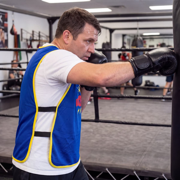 A man wearing a blue and yellow training vest throws a punch at a boxing bag in a gym setting.