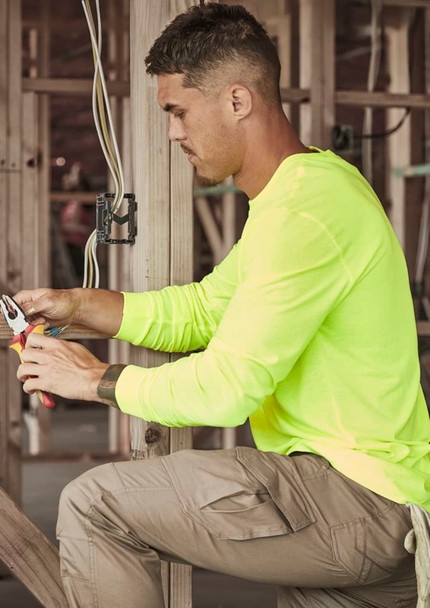 A man works in a construction setting, wearing a neon yellow long sleeve tee and beige cargo pants, using pliers.