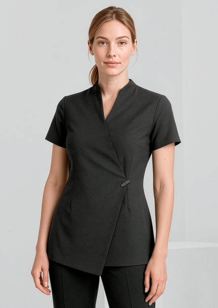 A woman is wearing a black spa tunic with a wrap design and short sleeves. The tunic features a simple button detail.